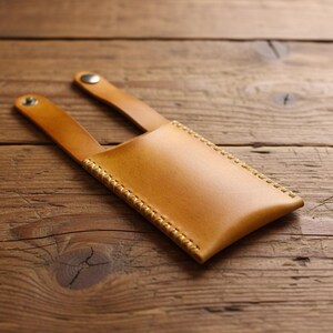 May include: A light brown leather phone case with a stitched border and two straps. The case is made of smooth leather and has a snap closure on the straps. The case is shown on a wooden surface.