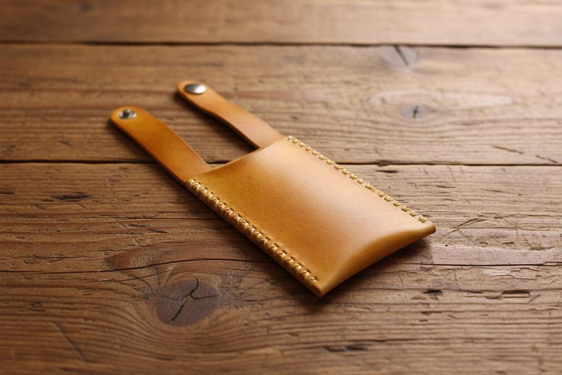 May include: A light brown leather phone case with a stitched border and two straps. The case is made of smooth leather and has a snap closure on the straps. The case is shown on a wooden surface.