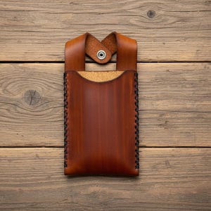 May include: A handcrafted, brown leather phone case with a loop and snap closure. The case features a stitched border and a cork interior. The leather has a rich, warm tone, and the case is set against a wooden background.