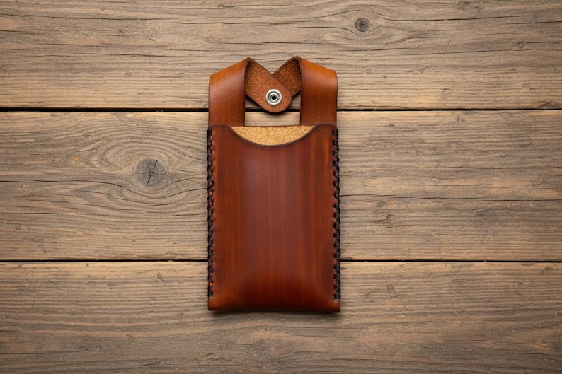 May include: A handcrafted, brown leather phone case with a loop and snap closure. The case features a stitched border and a cork interior. The leather has a rich, warm tone, and the case is set against a wooden background.
