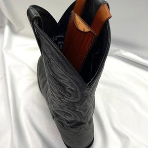 May include: A black leather cowboy boot with decorative stitching. Inside the boot is a brown leather wallet with a strap. The wallet is visible above the boot's opening, suggesting it's designed to be carried there.
