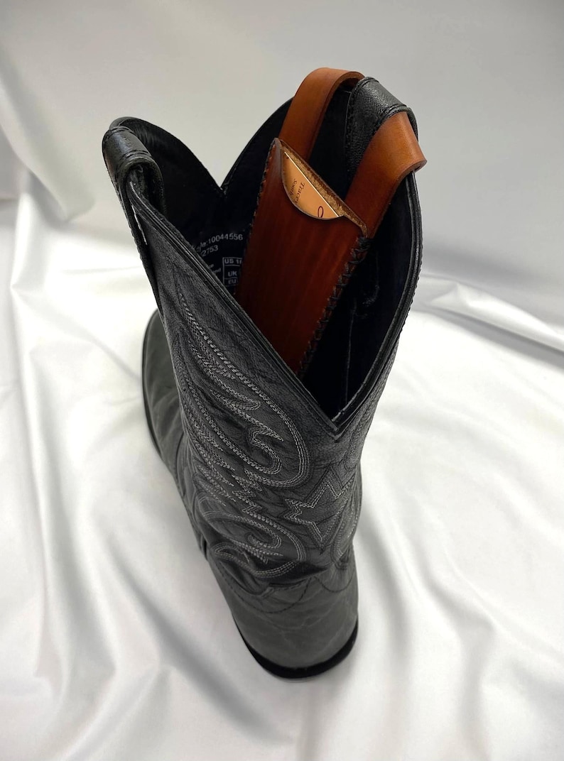 May include: A black leather cowboy boot with decorative stitching. Inside the boot is a brown leather wallet with a strap. The wallet is visible above the boot's opening, suggesting it's designed to be carried there.
