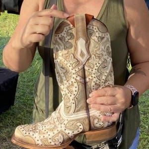 May include: A beige and white cowboy boot with intricate embossed patterns and studded accents. The boot features a square toe and a stacked heel. The upper part of the boot has a decorative design with a light brown leather trim.