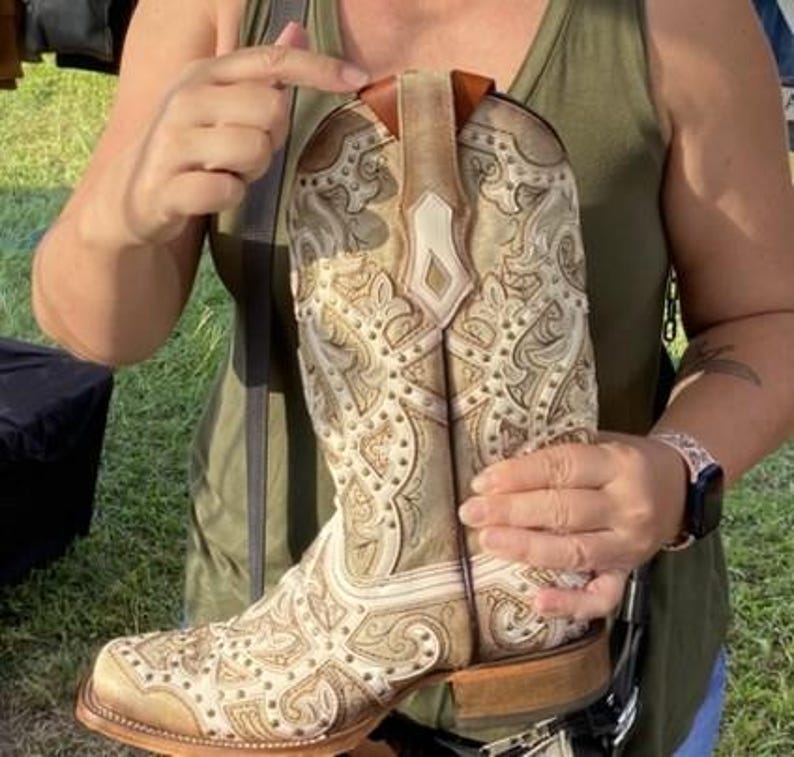 May include: A beige and white cowboy boot with intricate embossed patterns and studded accents. The boot features a square toe and a stacked heel. The upper part of the boot has a decorative design with a light brown leather trim.