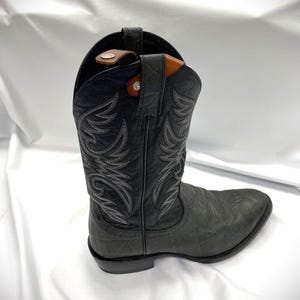May include: A black cowboy boot with decorative stitching in a lighter gray color. The boot has a textured finish and a pointed toe. A brown leather boot pull is visible at the top of the boot.