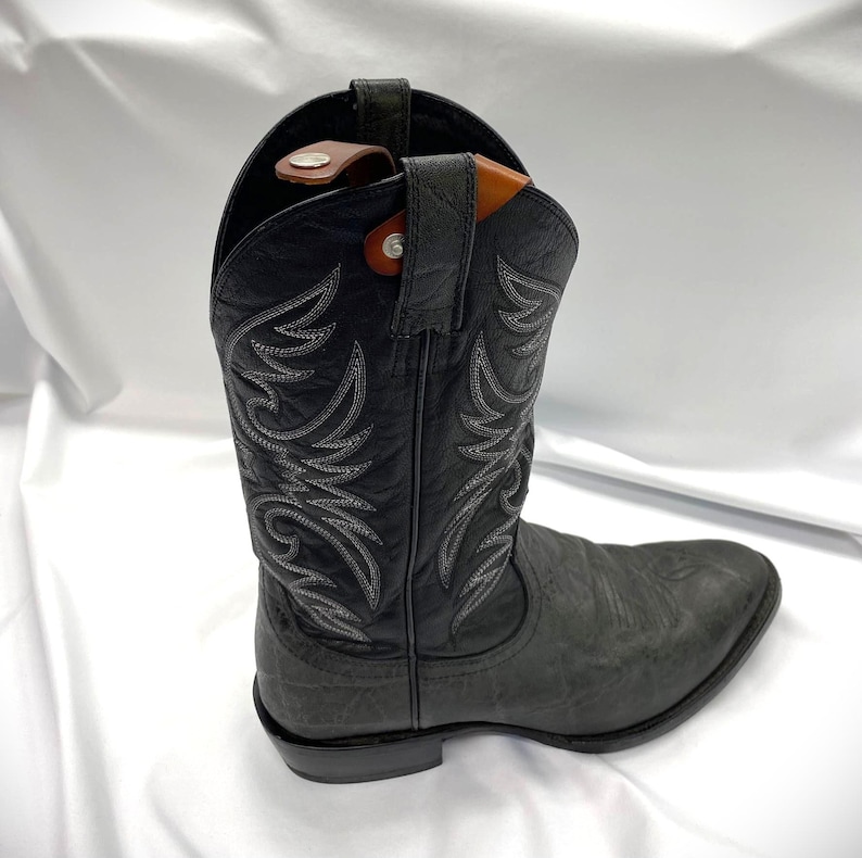 May include: A black cowboy boot with decorative stitching in a lighter gray color. The boot has a textured finish and a pointed toe. A brown leather boot pull is visible at the top of the boot.