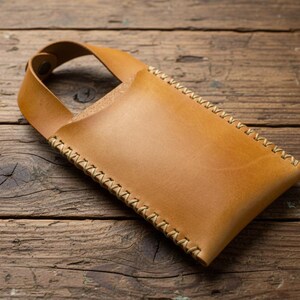 May include: A handcrafted, light brown leather phone case with a stitched edge and a handle. The case is rectangular with a curved top and is set against a rustic wooden background. The stitching is a contrasting light tan color.