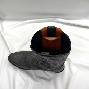 May include: A black leather cowboy boot with a brown and black leather wallet insert. The boot has decorative stitching and a pointed toe. The wallet is visible inside the boot's opening.
