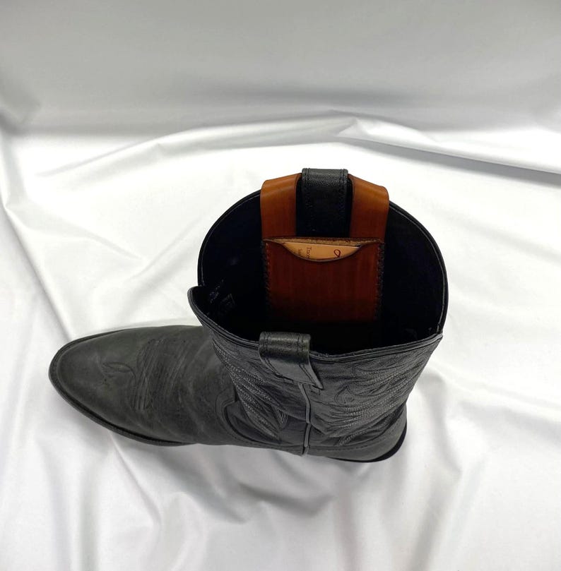 May include: A black leather cowboy boot with a brown and black leather wallet insert. The boot has decorative stitching and a pointed toe. The wallet is visible inside the boot's opening.
