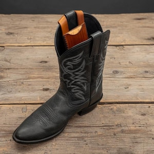 May include: A black leather cowboy boot with decorative stitching. A brown leather wallet is partially visible inside the boot. The boot is set on a rustic wooden surface.