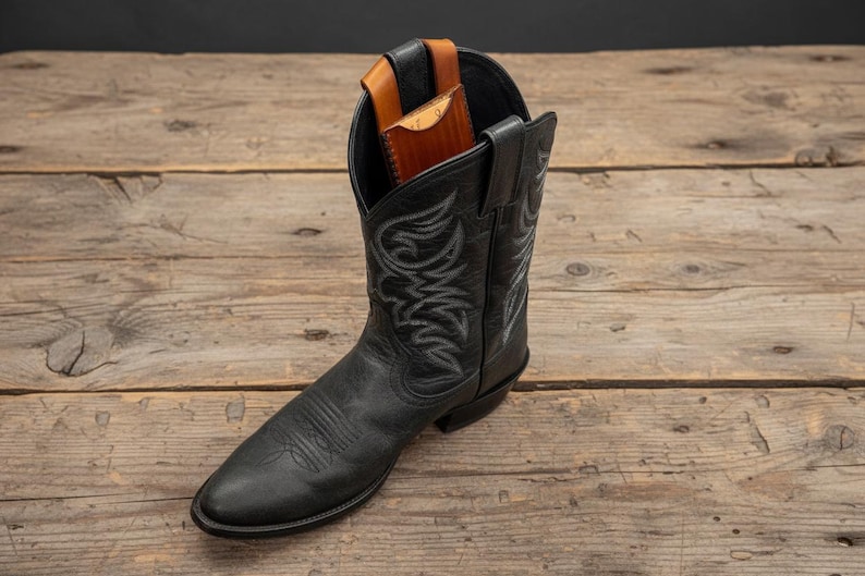 May include: A black leather cowboy boot with decorative stitching. A brown leather wallet is partially visible inside the boot. The boot is set on a rustic wooden surface.