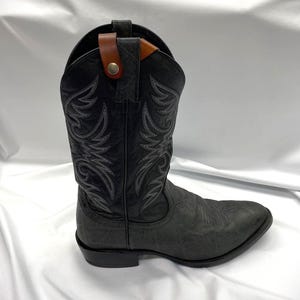 May include: A black cowboy boot with intricate white stitching and a textured finish. The boot features a brown leather strap with a button closure at the top. The boot has a pointed toe and a stacked heel.