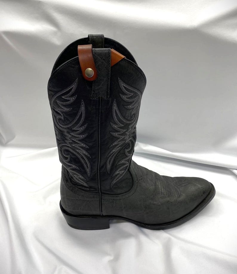 May include: A black cowboy boot with intricate white stitching and a textured finish. The boot features a brown leather strap with a button closure at the top. The boot has a pointed toe and a stacked heel.