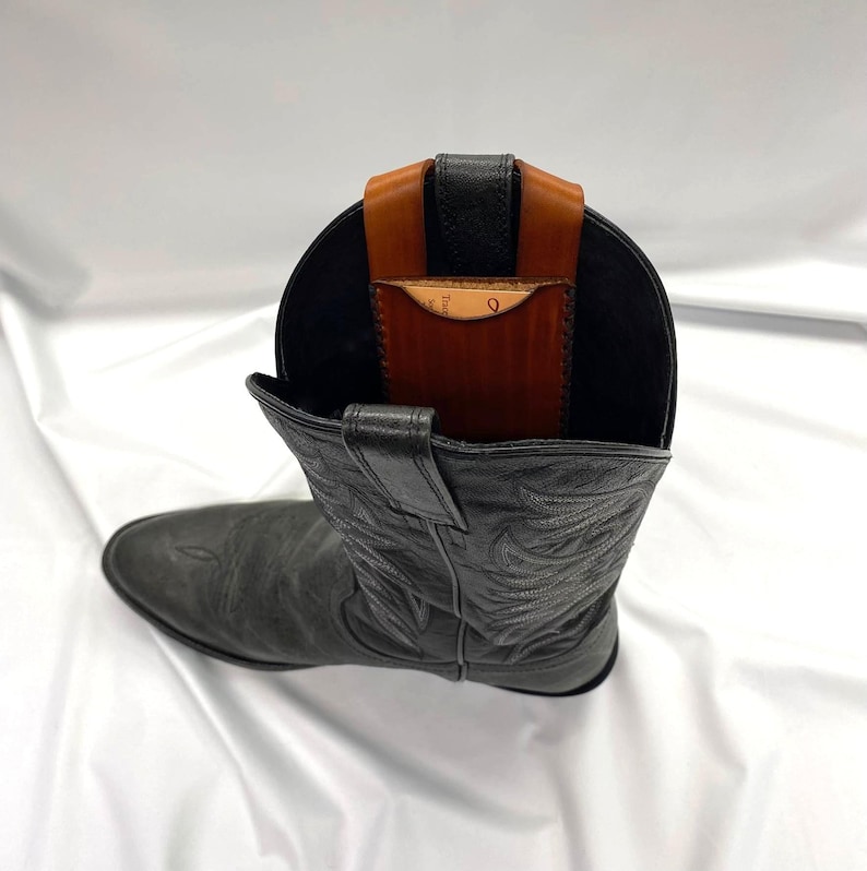 May include: A black leather cowboy boot with a brown and black leather wallet insert. The wallet is visible inside the boot, held in place by a strap. The boot is on a white surface.