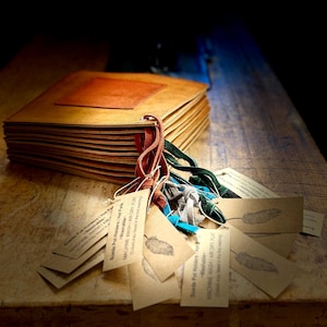 May include: A stack of handmade, light brown journals with a reddish-brown square on the cover. Attached are several tags with feather illustrations and text. The journals are on a wooden surface.