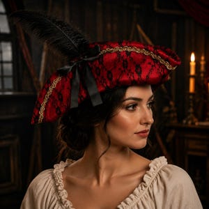 May include: A red tricorn hat featuring a black lace overlay, gold braided trim along the edges, and a black satin ribbon bow. A tall, dark black feather plume is attached to the side of the hat, which is worn over a beige ruffled shirt.