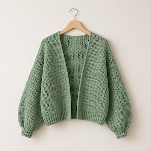 Può includere: Un cardigan aperto color verde salvia caratterizzato da un motivo a uncinetto testurizzato, maniche lunghe a palloncino con polsini a coste e un orlo a coste. Il capo è appeso a una gruccia in legno e presenta una lavorazione uniforme.