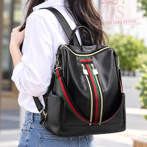 May include: A black backpack featuring a vertical stripe pattern in red and green down the center, gold-tone metal zippers, and a detachable shoulder strap with matching red and green stripes. The bag has a smooth finish and side pockets.