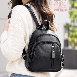 May include: A small black backpack with a textured, pebbled finish, featuring a main zippered compartment, a smaller front zippered pocket with a vertical pull tab, and adjustable shoulder straps. The logo Softie Land Design is visible.
