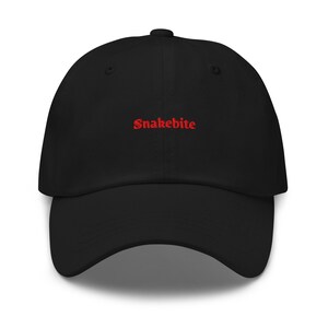 May include: A black baseball cap featuring the word Snakebite embroidered in red thread across the front centre. The cap has a structured crown, a curved brim, and matching black eyelets for ventilation, creating a clean and simple design.