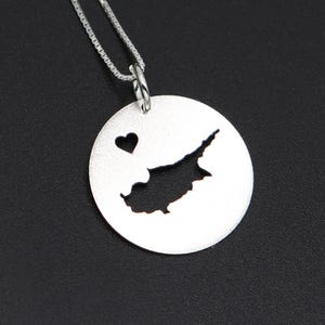 Op de afbeelding: Een zilveren hangerketting met een ronde bedel met een uitsparing van het eiland Cyprus en een hart. De hanger hangt aan een fijne zilveren ketting, tegen een donkere achtergrond. Het is een sieraad.