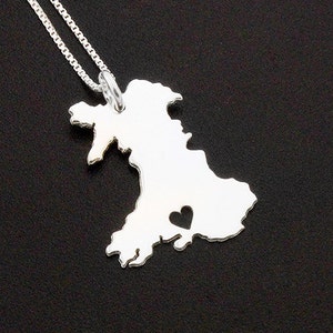 May include: A silver pendant necklace depicting the shape of Wales, with a heart-shaped cutout. The pendant is suspended from a fine silver chain. The necklace is shown against a dark backdrop.