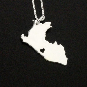 May include: A silver necklace featuring a pendant shaped like a country's outline, with a heart-shaped cutout. The pendant hangs from a delicate chain, creating a simple yet meaningful piece of jewelry. The necklace is displayed against a dark background.