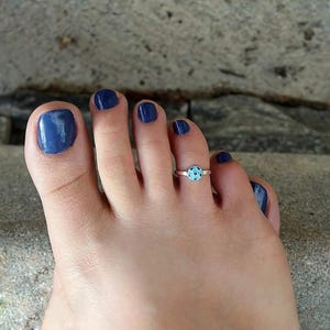 May include: A silver toe ring featuring a small, circular charm with a light blue background and dark blue polka dots. The ring is worn on the second toe, which is positioned next to a large toe with dark blue nail polish on all visible nails.