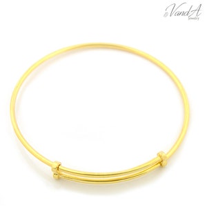 May include: A gold-toned expandable bangle bracelet featuring a smooth, thin circular wire design. The bracelet includes two small sliding metal connectors that allow for size adjustment, with the text VandA Jewelry visible in the top right corner.