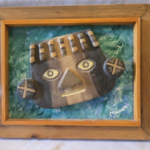 Può includere: Un'opera d'arte da parete incorniciata che presenta una maschera in ceramica marrone scuro tridimensionale con tratti del viso stilizzati, inclusi occhi a mandorla e orecchie circolari con segni a croce, su uno sfondo dipinto blu e verde.