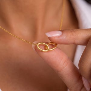 Könnte beinhalten: Eine goldfarbene Halskette mit zwei kleinen, ineinandergreifenden ovalen Gliedern als zentraler Anhänger. Die Kette ist dünn und zart und mit beiden Seiten der ovalen Glieder verbunden. Das Metall hat eine glatte, polierte Oberfläche in einem warmen Gelbton.