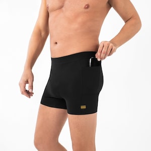 Puede incluir: Pantalones cortos de compresión atléticos negros con una cintura ancha y un bolsillo lateral en la cadera derecha. El bolsillo contiene un pequeño dispositivo electrónico negro. Se ve una pequeña etiqueta rectangular amarilla con texto negro.
