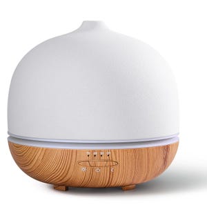 May include: A dome-shaped essential oil diffuser with a matte white top and a light brown wood grain base. The base features three small feet, a horizontal control panel with labels 1H, 2H, 3H, ON, and three icons representing light, power, and mist.