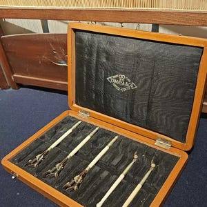 Könnte beinhalten: Ein technisches Zeicheninstrumenten-Set aus Holz in einem rechteckigen Koffer mit schwarzem Samt-Innenfutter. Das Set enthält fünf Werkzeuge aus Metall und cremefarbenem Material. Der Innendeckel zeigt das Logo Tamaya & Co Ginza Tokyo.