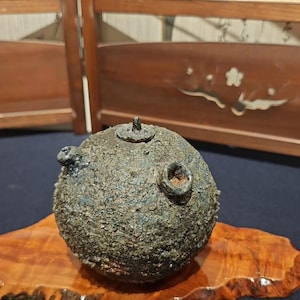 May include: A spherical, dark-toned ceramic vessel with a heavily textured, rough surface. The object features a small, centred lid with a knob and two additional circular openings on its upper portion, showcasing a weathered, earthy appearance.