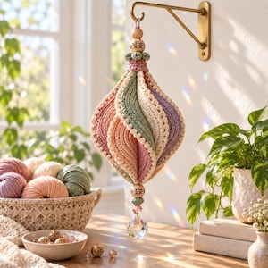 Könnte beinhalten: Ein hängendes Häkelornament mit spiralförmigen Bahnen in gedeckten Tönen von Rosa, Salbeigrün, Creme und Lavendel. Das Stück ist mit Holzperlen, kleinen grünen Häkeldetails und einem klaren, facettierten Kristallprisma am unteren Ende verziert.
