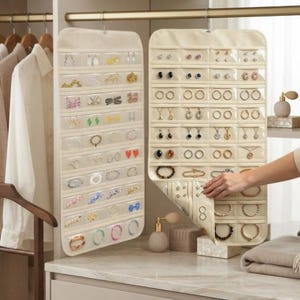 May include: Two hanging jewelry organizers made of cream-colored fabric with multiple clear plastic pockets. Each organizer features rows of transparent slots designed to hold various earrings, bracelets, and necklaces, suspended from a metal rod.