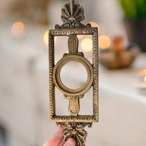 May include: A rectangular brass-colored metal plate featuring an ornate border with a central circular opening. The piece includes decorative shell-like motifs at both the top and bottom ends, with textured detailing along the frame edges.