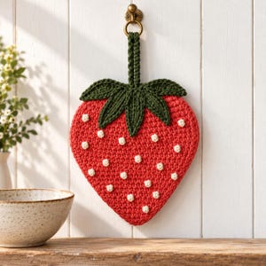 Può includere: Una presina all'uncinetto a forma di fragola, caratterizzata da un corpo rosso vivace con piccoli punti bianchi testurizzati, sormontata da foglie all'uncinetto verde scuro e un occhiello per appenderla, sospesa a un gancio da parete color ottone.