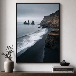 Op de afbeelding: Een ingelijste wandkunstprint met een dramatisch kustlandschap met een zwart zandstrand, donkere oceaangolven en torenhoge bruine kliffen onder een bewolkte grijze lucht. In de verte steken grillige rotsformaties uit het zeewater.