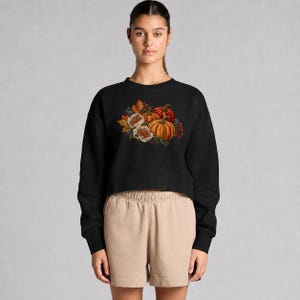May include: A black long-sleeve cropped sweatshirt featuring a central graphic of orange pumpkins, white flowers, and autumn leaves. The sweatshirt is paired with beige shorts that have a visible elastic waistband and a straight-leg cut.