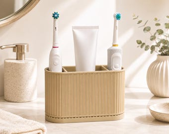 May include: A beige, oval-shaped bathroom organiser with a vertical ribbed texture. The container features four distinct compartments, currently holding two white electric toothbrushes and one blank white tube of toothpaste in an upright position.