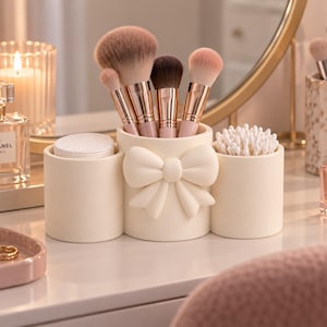 Puede incluir: Un organizador de tocador de color crema con tres compartimentos y un lazo decorativo en la sección central. El organizador contiene cuatro brochas de maquillaje con virolas de oro rosa, discos de algodón a la izquierda y bastoncillos a la derecha.