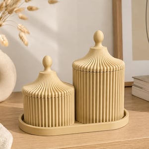 May include: Two beige cylindrical storage jars with vertical ribbed textures and matching lids topped with spherical finials. The jars are of different heights and sit together on a matching oval-shaped tray, creating a cohesive set.