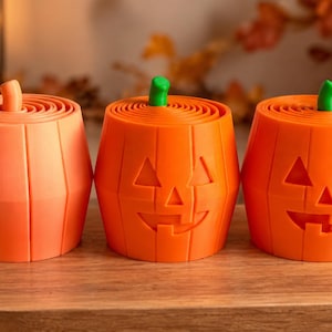 May include: Three orange pumpkin-shaped containers with green stems. The left container has a smooth exterior, while the two on the right feature carved jack-o'-lantern faces with triangular eyes and noses and wide, toothy grins. Each has a spiral top.