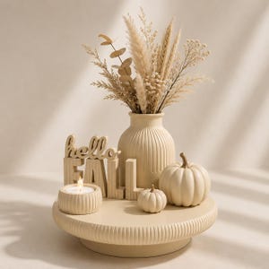 May include: A beige decorative set featuring a ribbed vase filled with dried grasses, a round ribbed candle holder with a lit tea light, two smooth pumpkins, and a block sign with the text "hello FALL" all arranged on a matching ribbed pedestal.