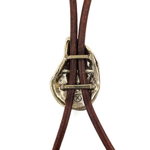 May include: A gold-toned bolo tie with a unique, abstract design. The tie features a brown leather cord and a metal clasp with a cross-shaped design.