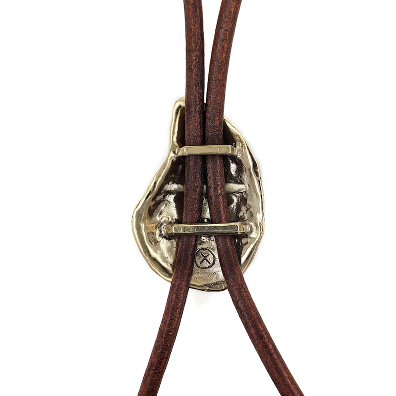 May include: A gold-toned bolo tie with a unique, abstract design. The tie features a brown leather cord and a metal clasp with a cross-shaped design.