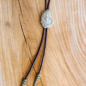 May include: A brown leather bolo tie with a silver, textured, oval-shaped pendant. The tie has two decorative tips.