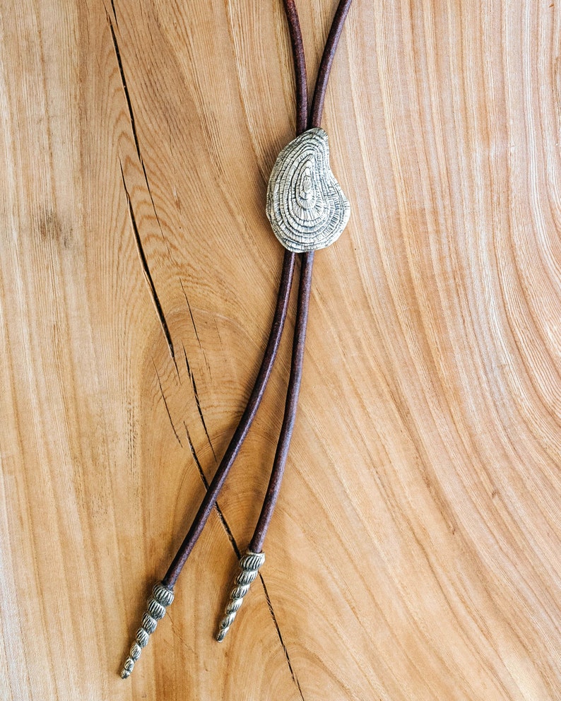May include: A brown leather bolo tie with a silver, textured, oval-shaped pendant. The tie has two decorative tips.